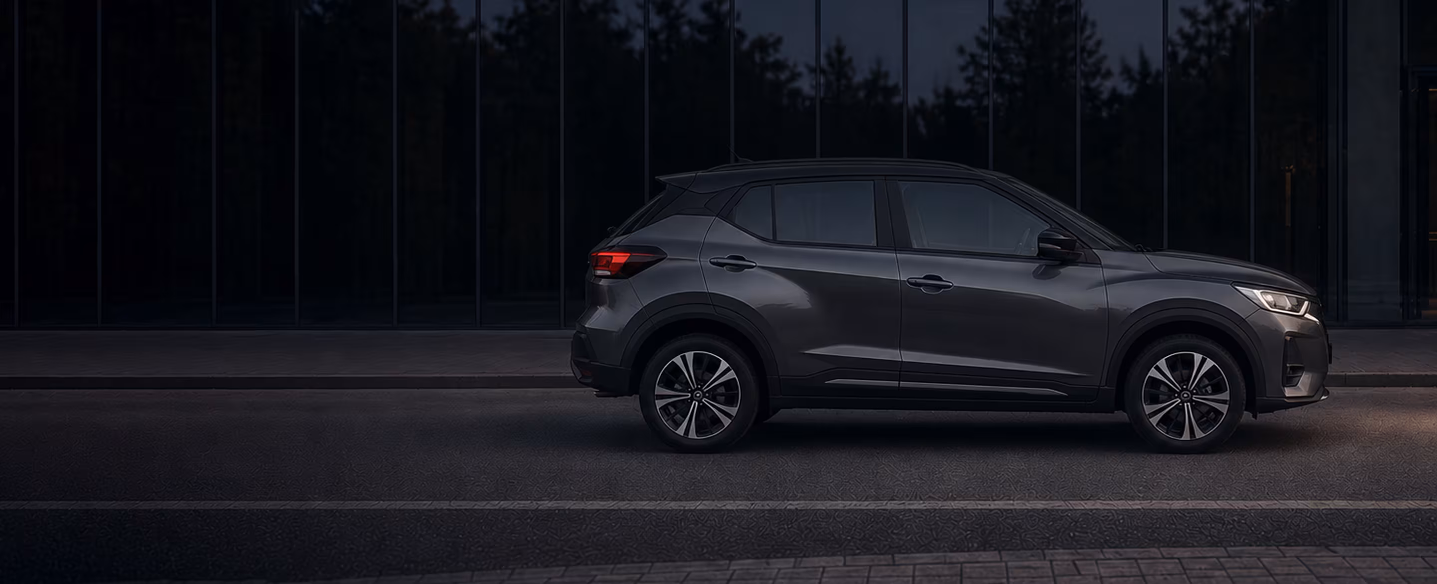 Dark gray compact hatchback parked on a dimly lit street in front of reflective glass windows.