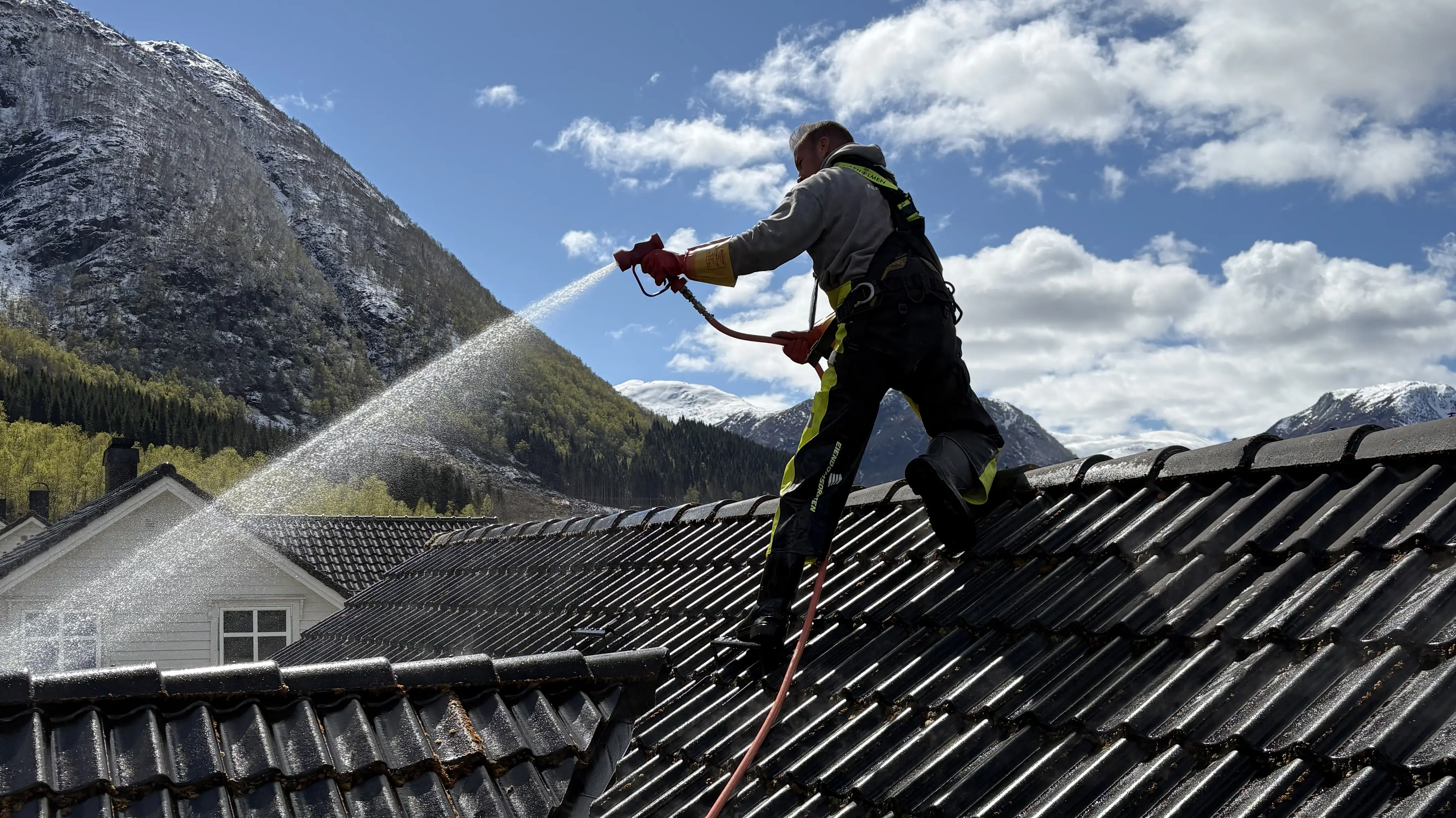 Mann i arbeid med å spyle tak med vann i fjellområde under blå himmel med skyer.