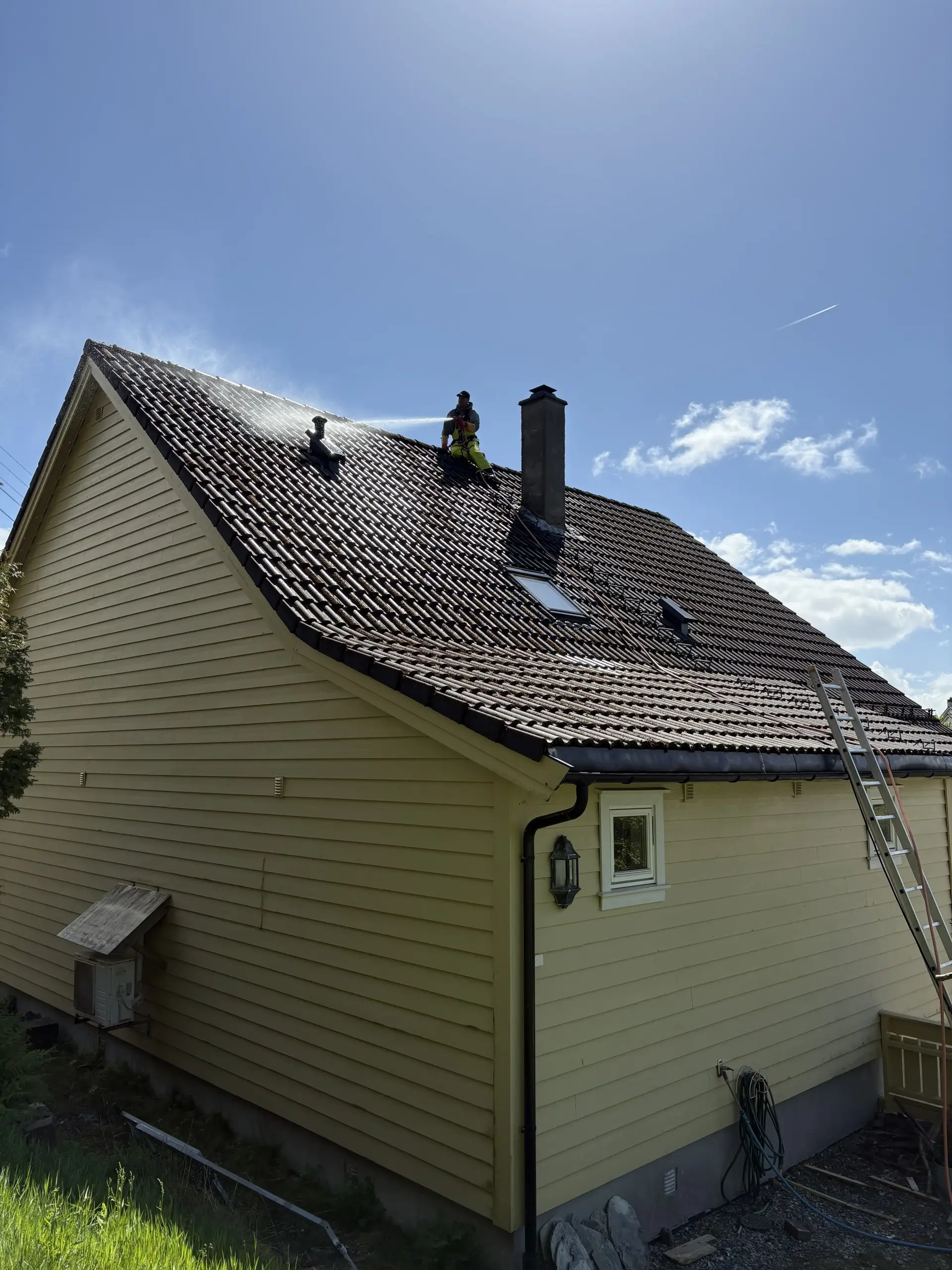 Person rengjør tak med høytrykksspyler på et gult hus under en blå himmel.