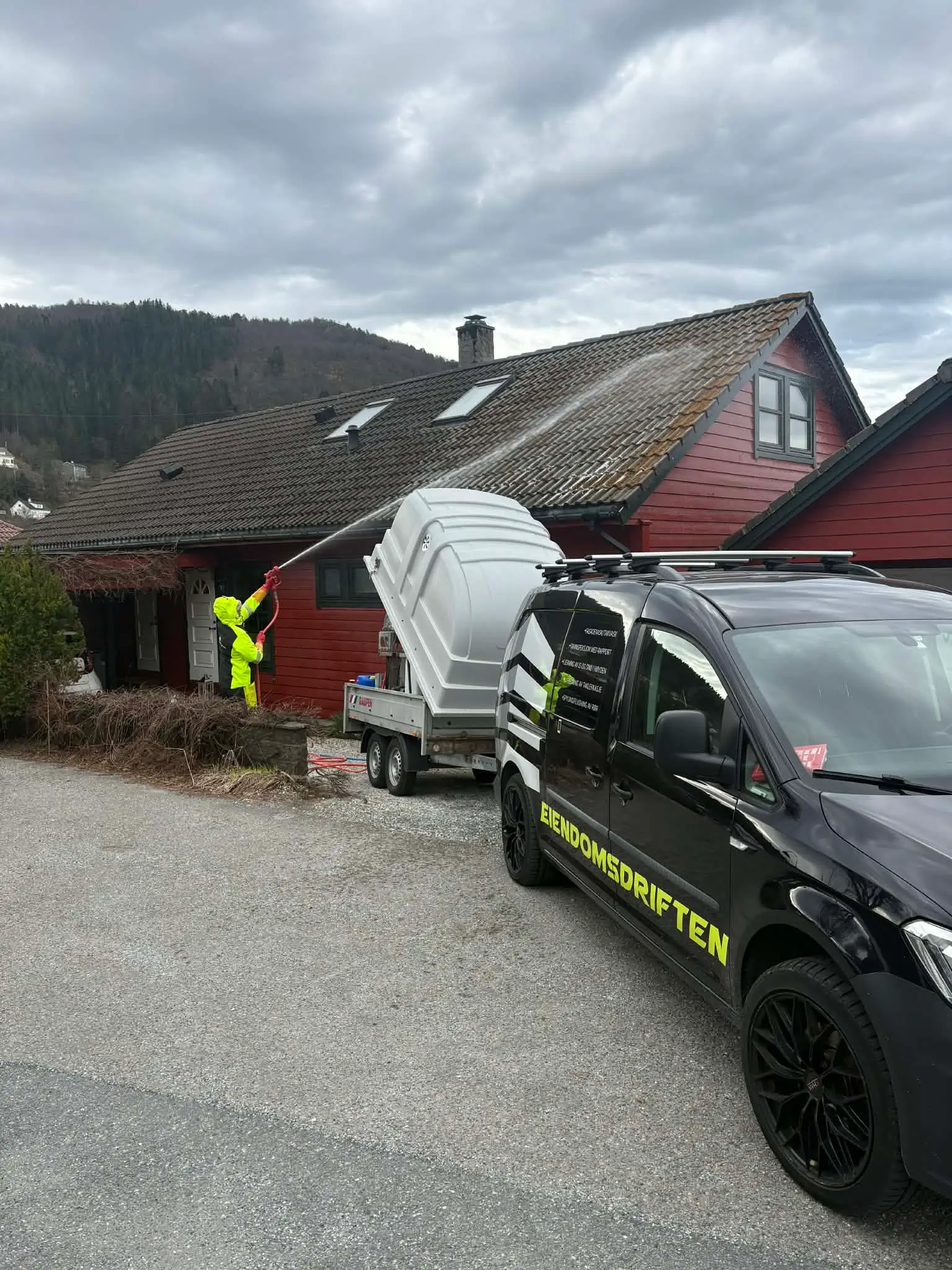 Person i gul arbeidsdress rengjør taket på rødt hus med høytrykksspyler fra biltilhenger.