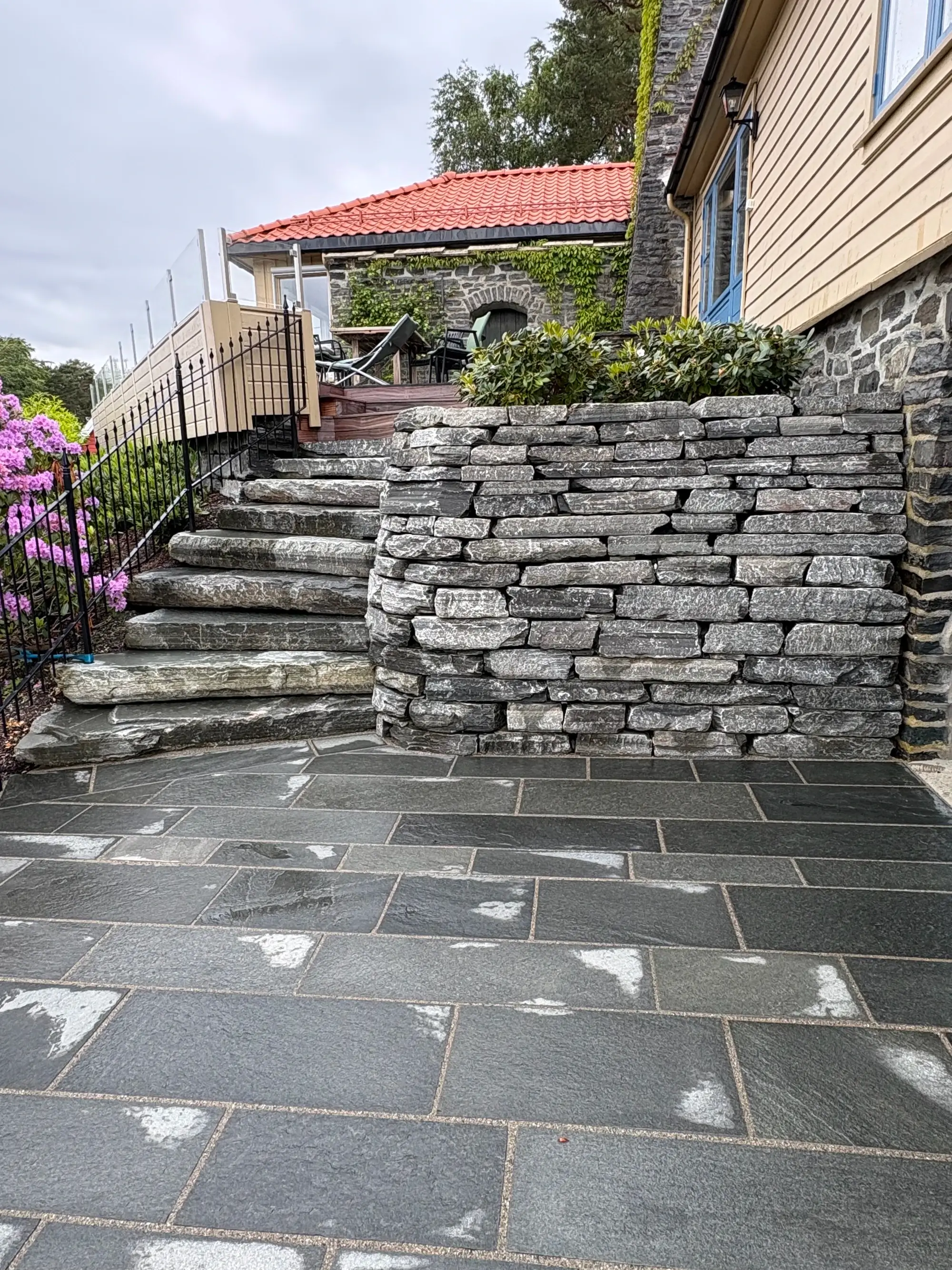 Steintrapp og belegningsstein ved hus med rødt tak og steinmur med planter.
