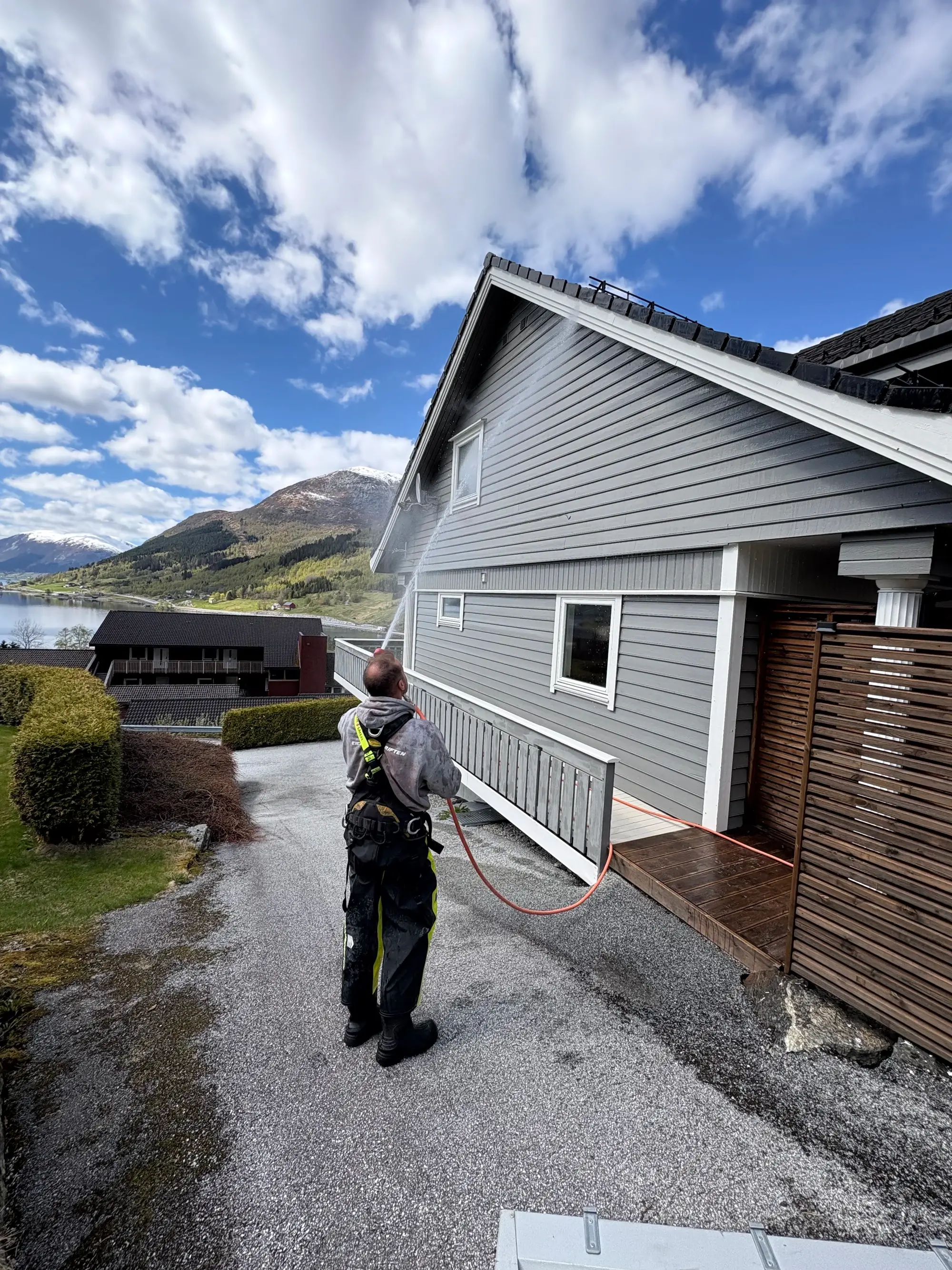 Person som høytrykksspuler en grå husfasade på en gårdsplass med fjell og blå himmel i bakgrunnen.