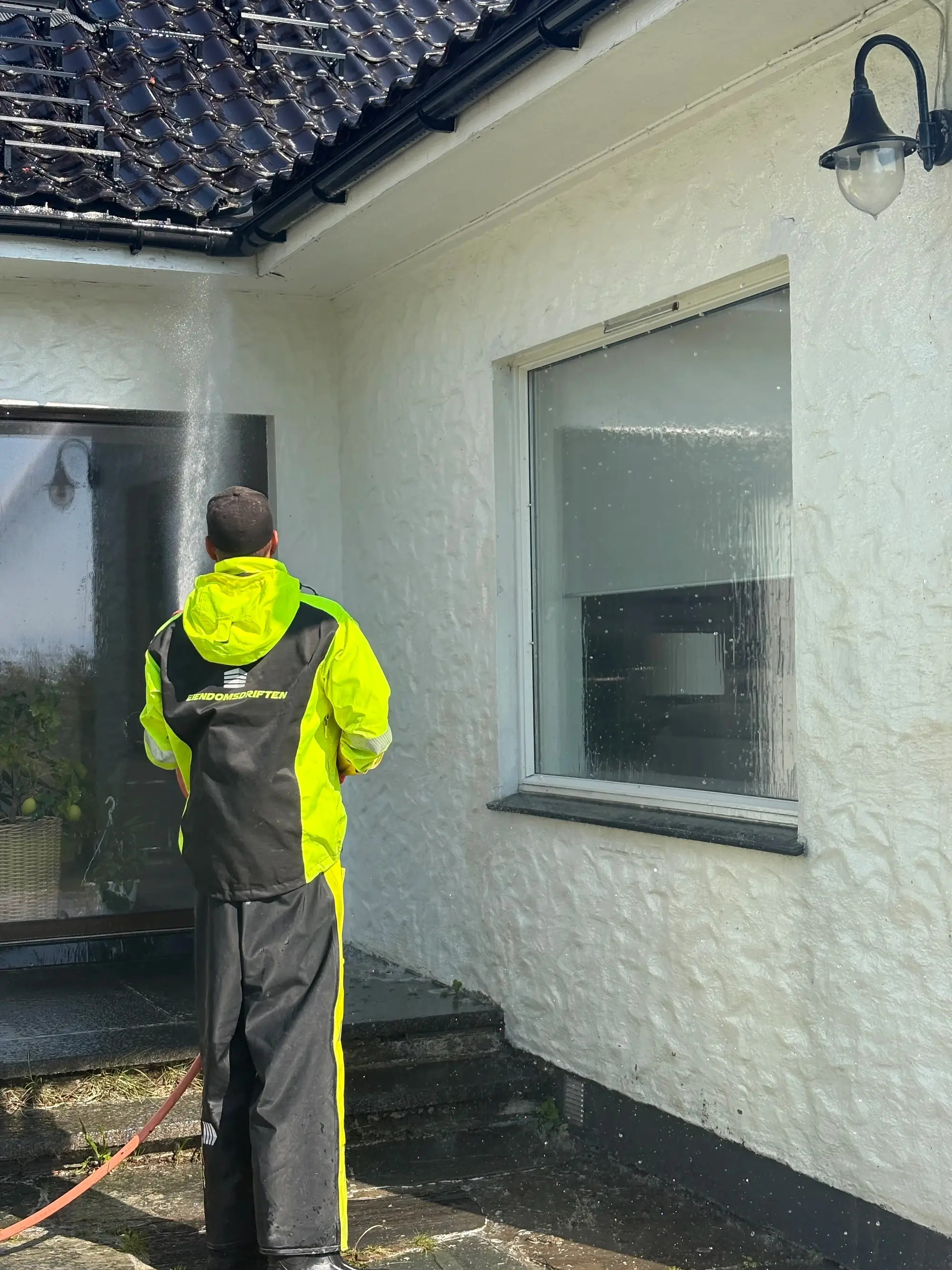 Person i verneklær høytrykksspuler en hvit fasade på et hus utendørs.