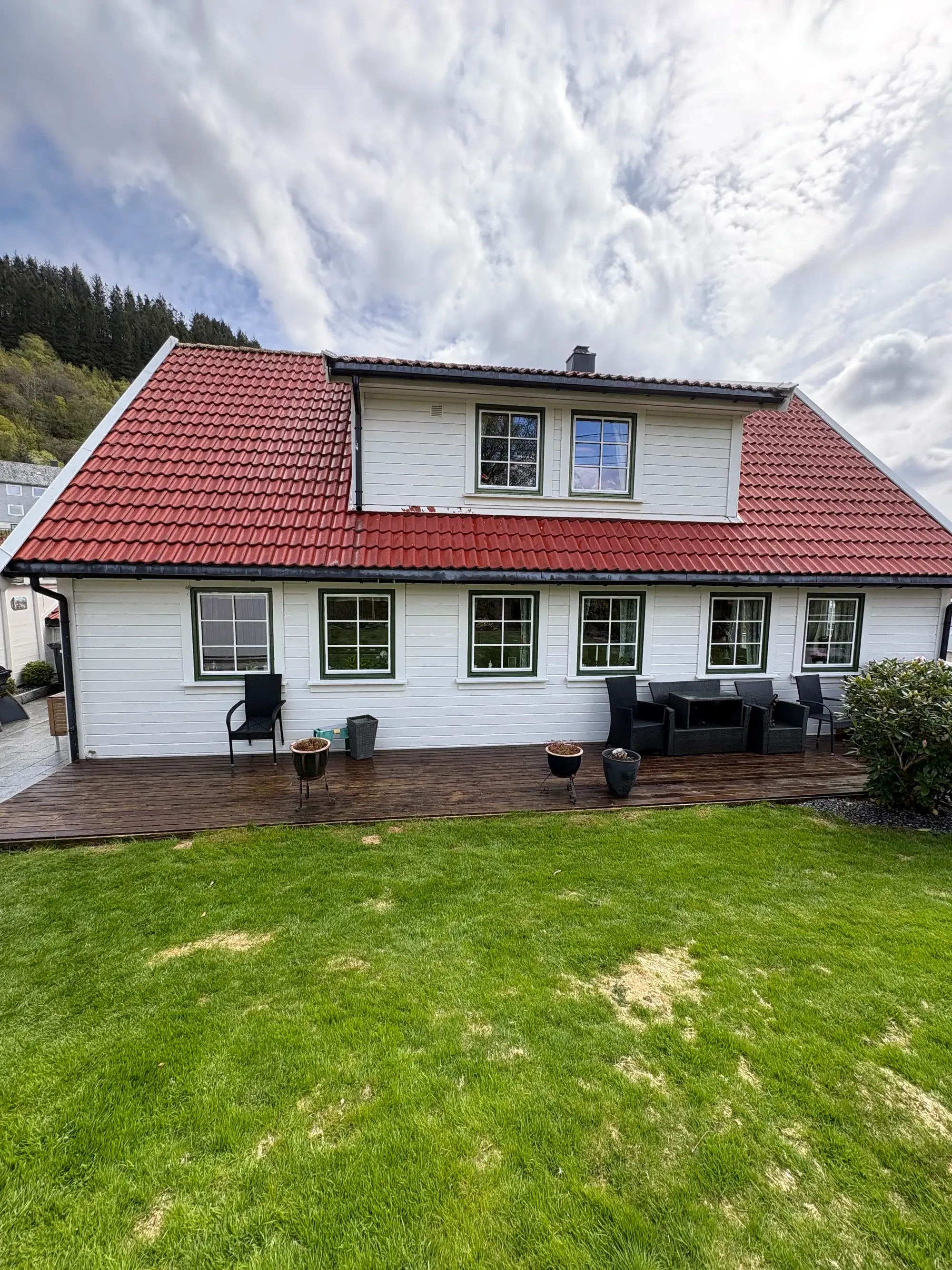 Hvit to-etasjes bolig med rødt tak og veranda med utemøbler og blomsterpotter foran en gressplen.
