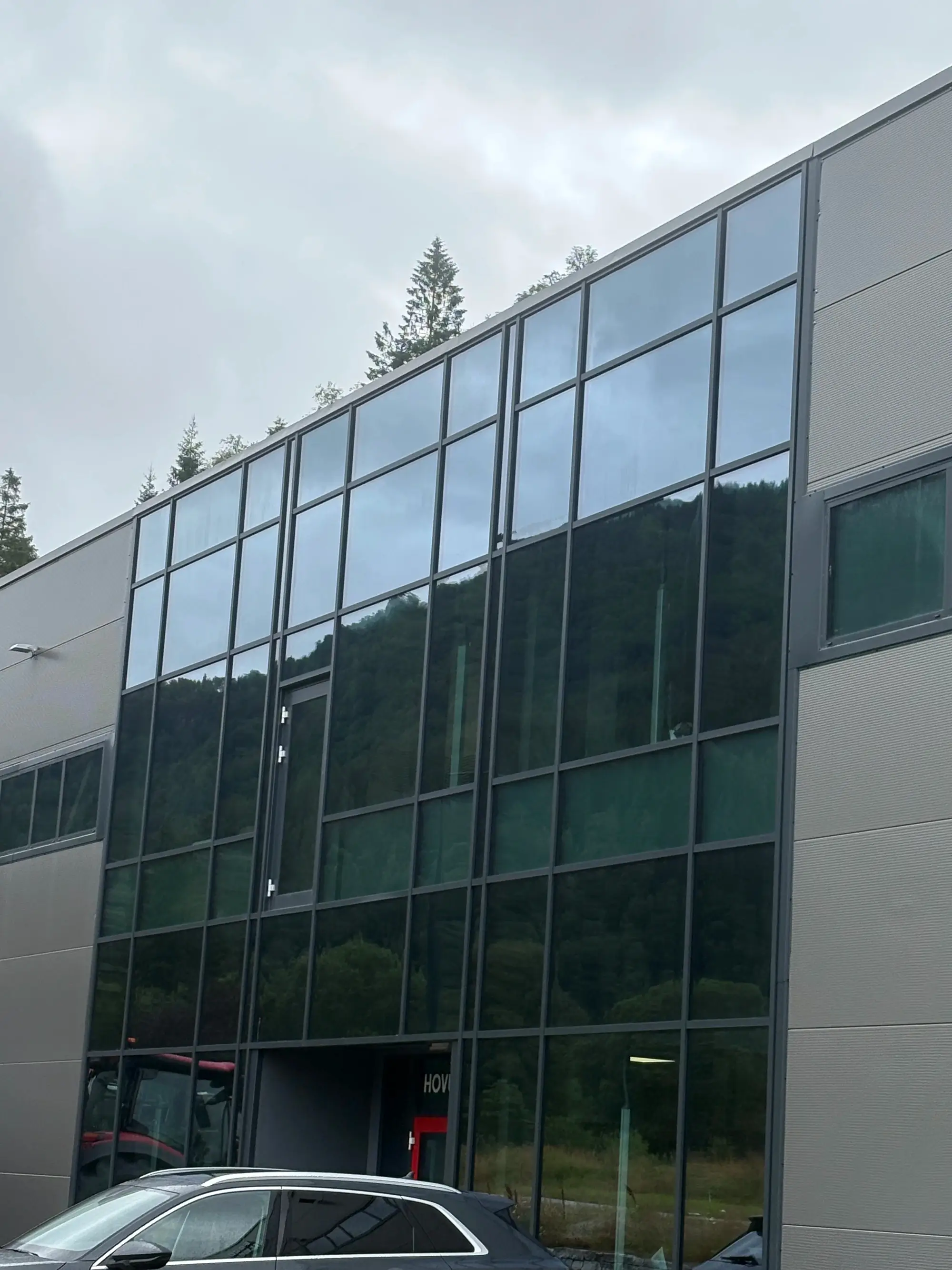 Moderne glasfasade på en bygning med bil parkert foran og trær i bakgrunnen.