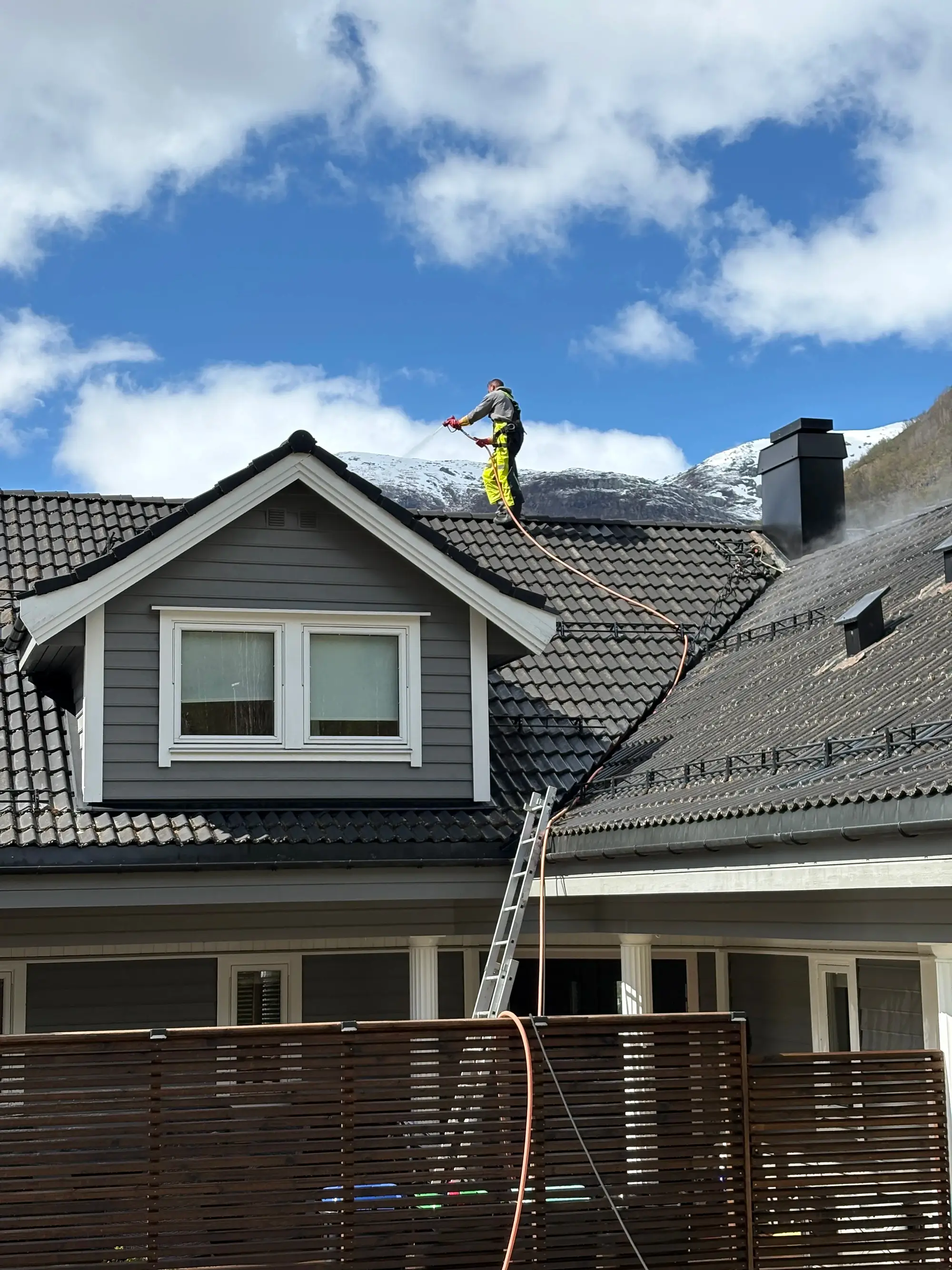 Mann vasker tak med høytrykkssprøyte på grått hus med svarte takfliser under blå himmel.