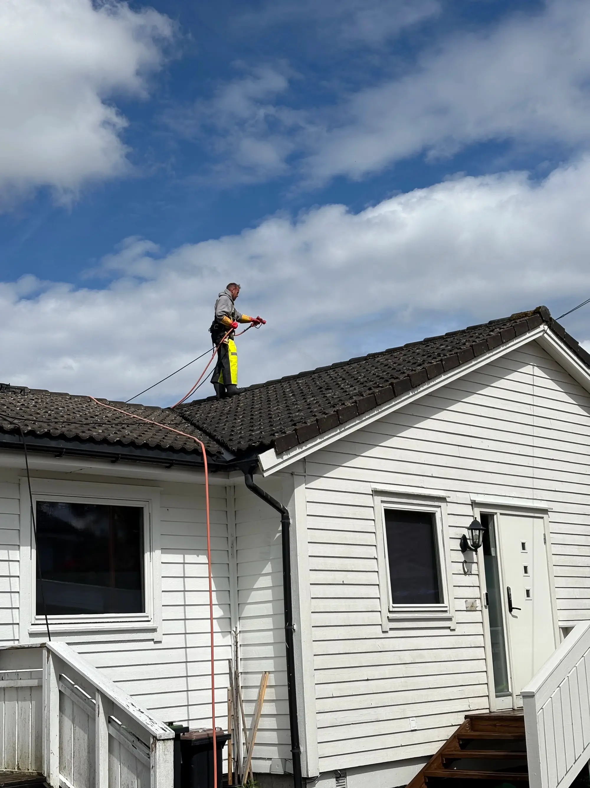 Person on roof pressure washing dark roof tiles on a white wooden house under cloudy blue sky.