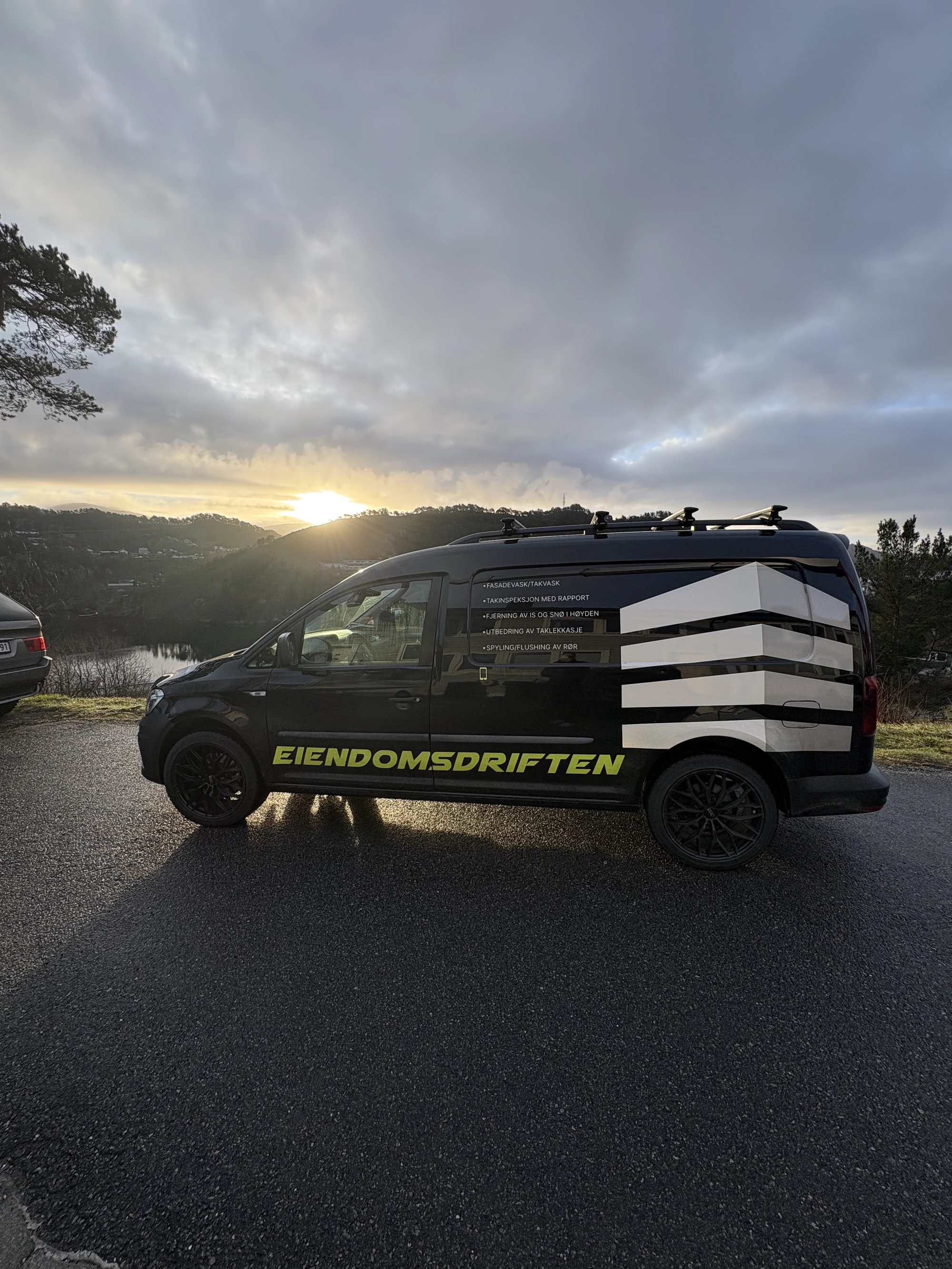 Svart varebil med teksten EIENDOMSDRIFTEN parkert i solnedgang ved vann og trær.
