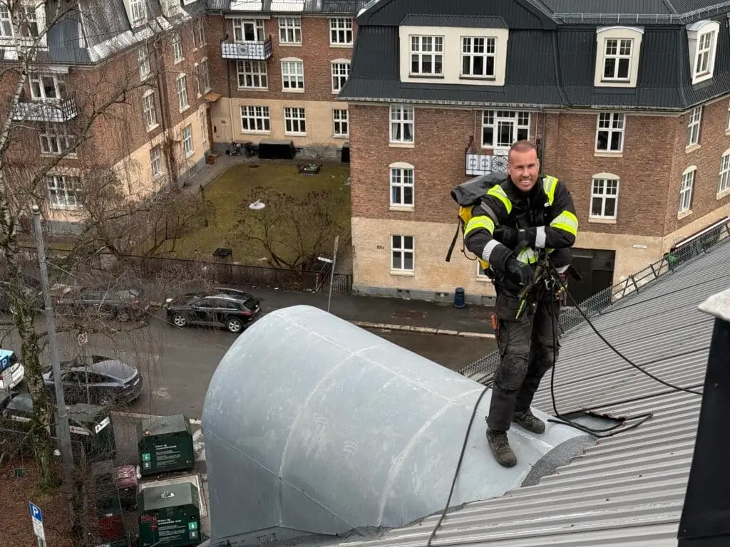 Arbeider i sikkerhetsutstyr på tak i urbant område med murbygninger og biler i bakgrunnen.
