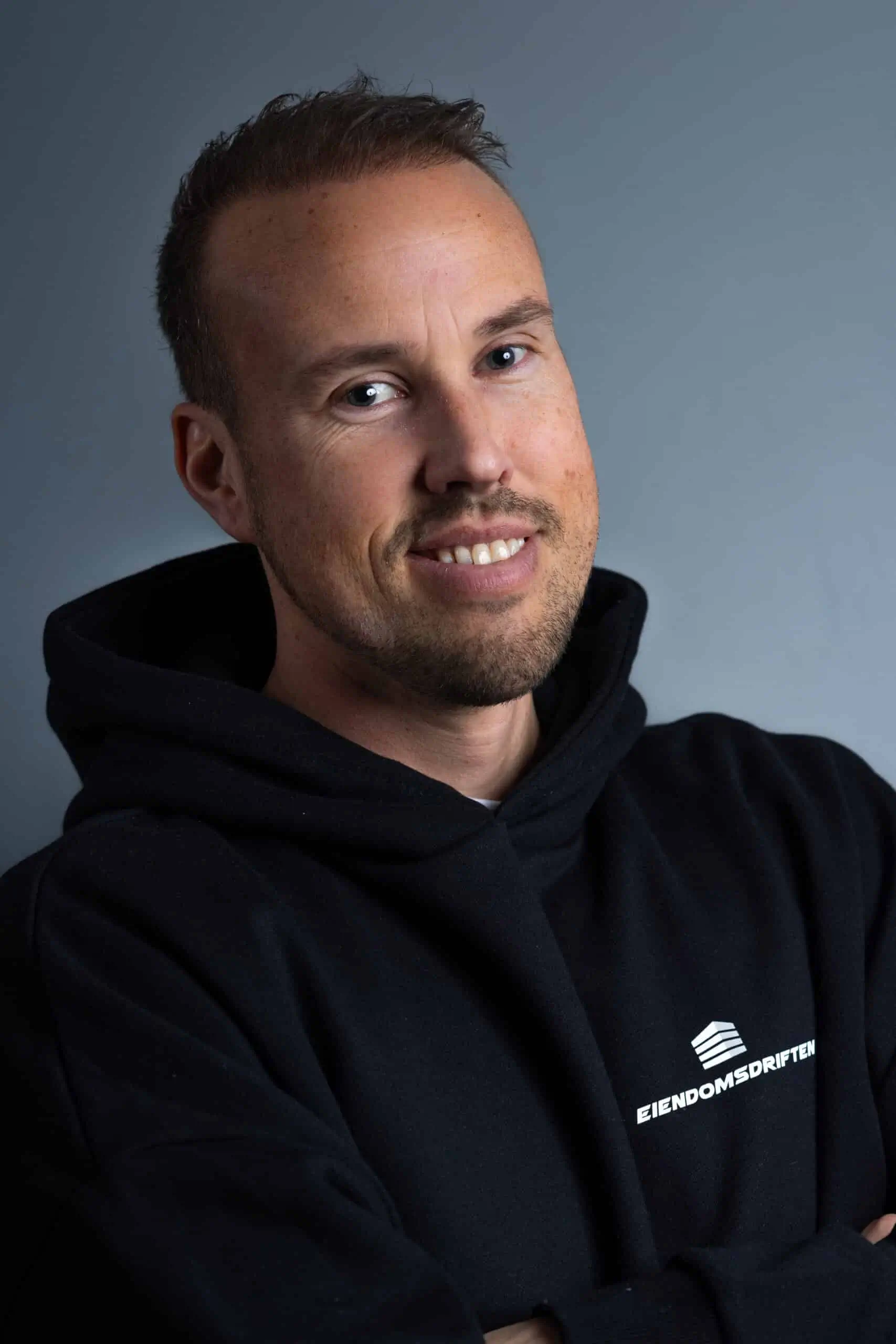 Portrett av en smilende mann i svart hettegenser med teksten Eiendomsdriften.