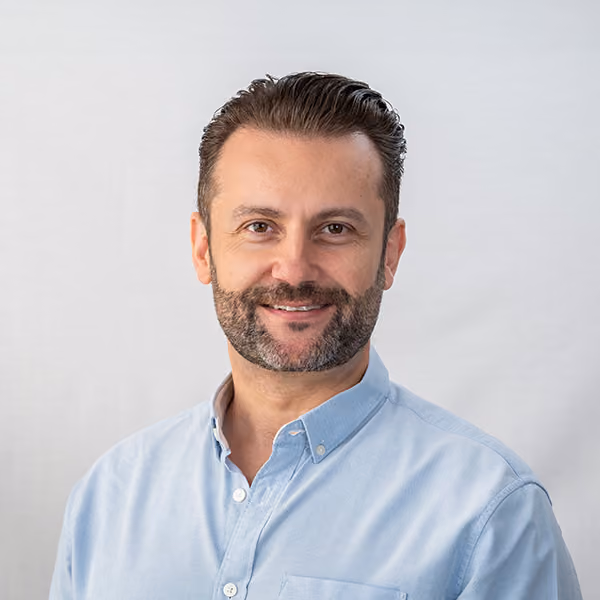 Smiling man with slicked-back hair and beard wearing a light blue button-up shirt against a plain background.