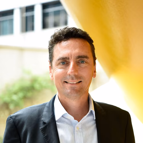Man with short dark hair wearing a black blazer and white shirt, smiling outdoors by a yellow structure.