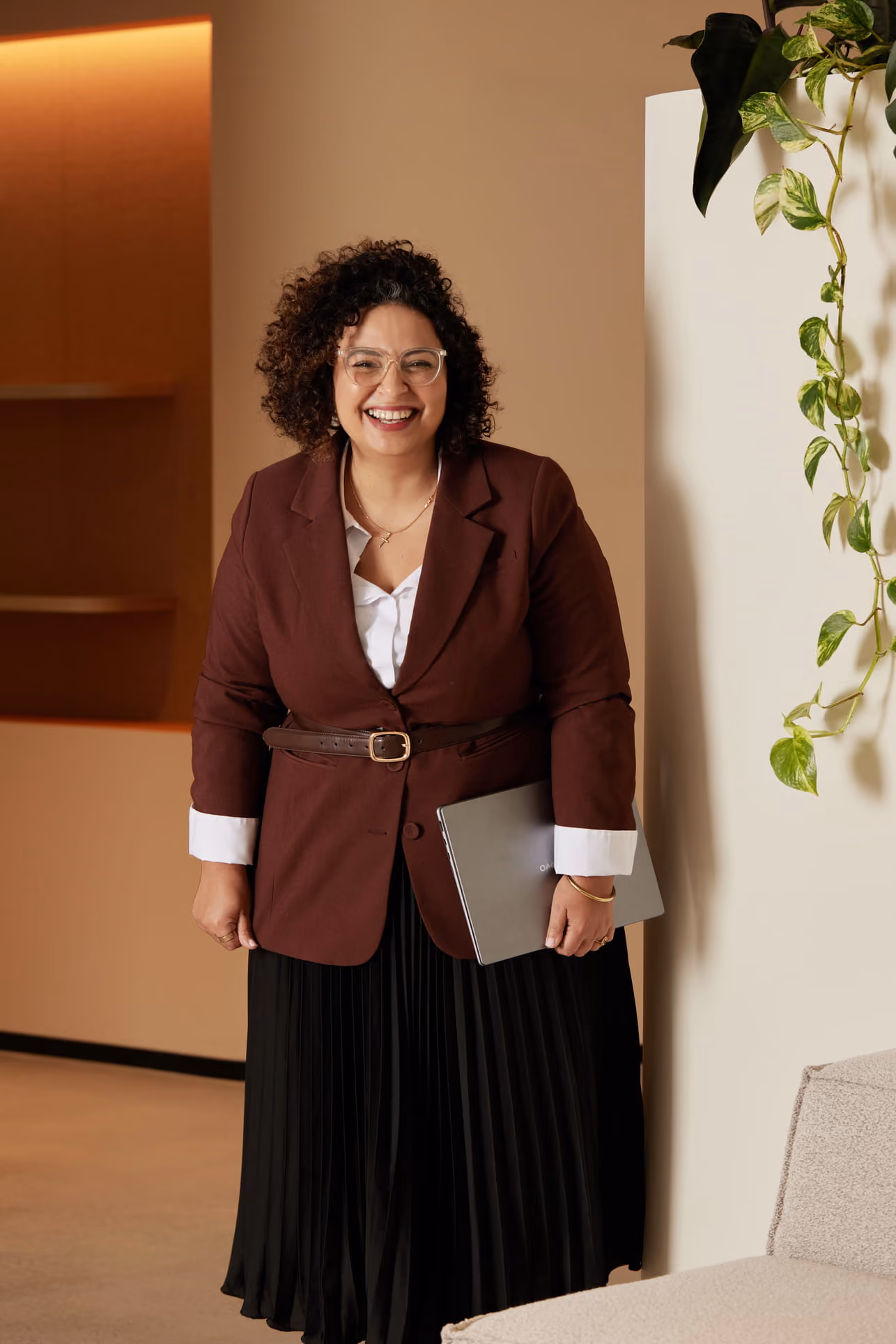 Smiling woman with curly hair wearing glasses, brown blazer, and black skirt, holding a laptop.