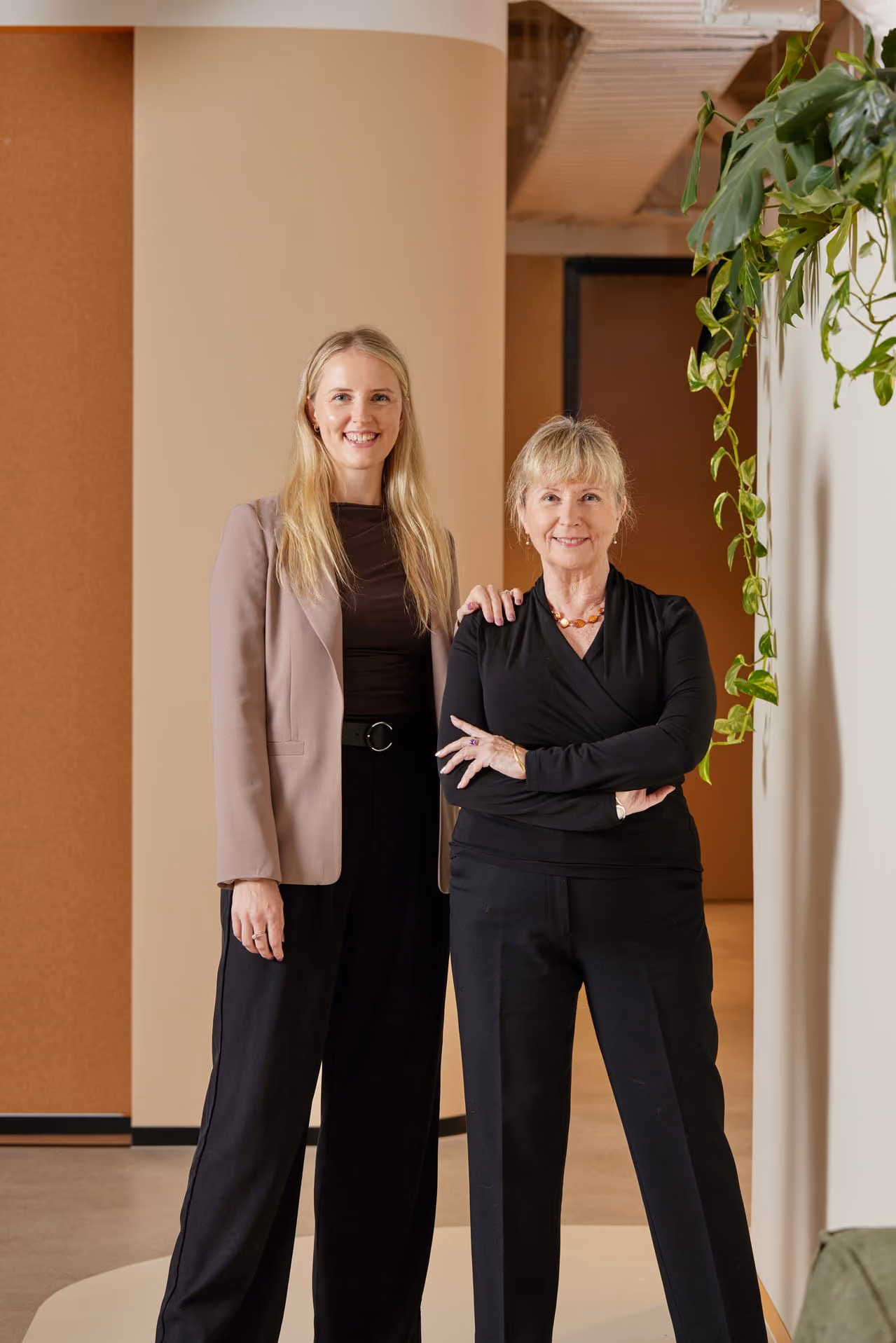 Two women smiling, standing indoors next to each other with one woman’s hand on the other's shoulder.