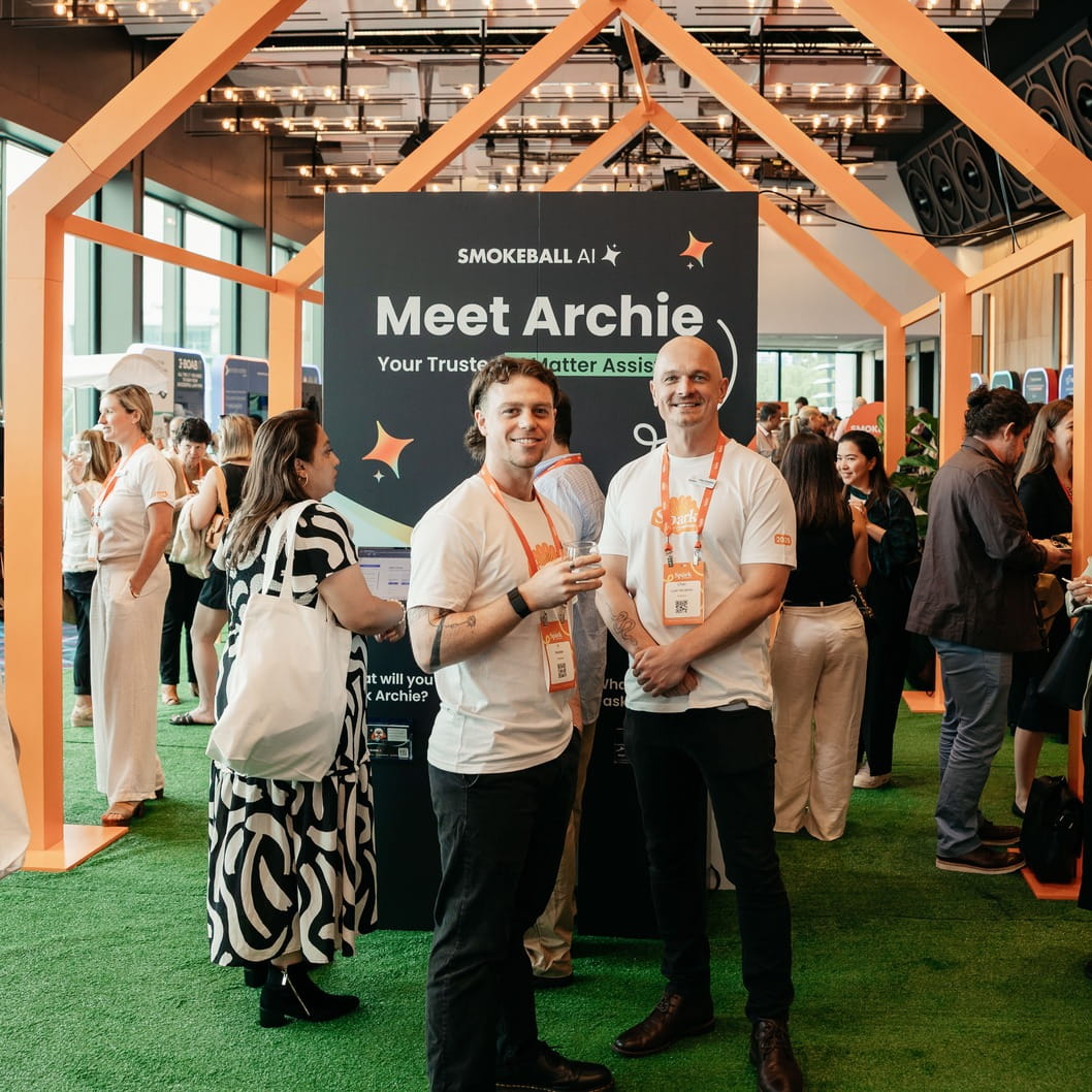 Two men in white t-shirts with lanyards stand in front of a Smokeball AI Meet Archie booth at an event.