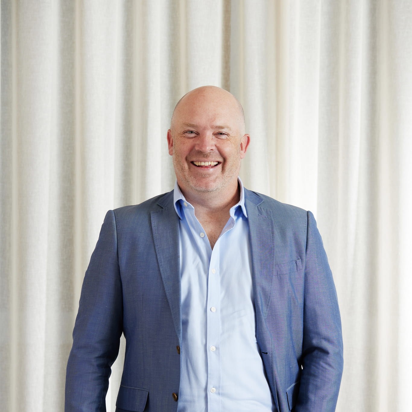 Middle-aged bald man smiling, wearing a blue blazer and light blue shirt, standing before cream curtains.