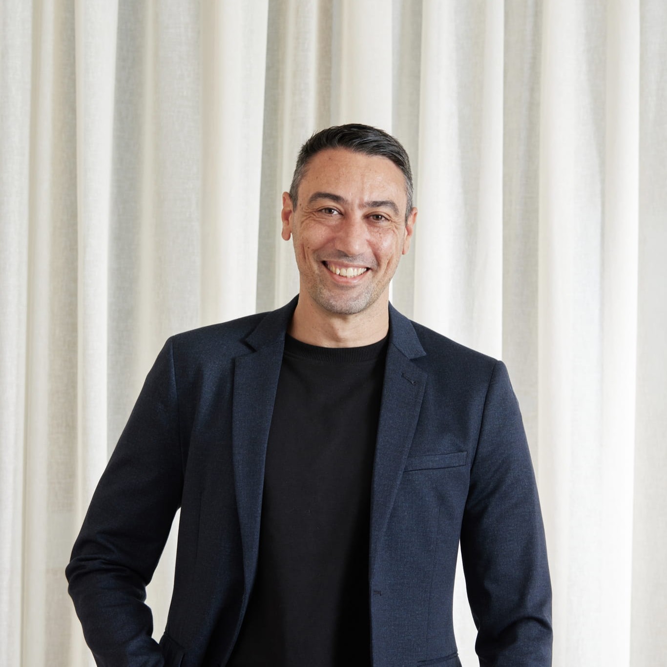 Smiling man with short dark hair wearing a dark blazer and black shirt in front of light curtains.