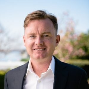 Smiling man with short light brown hair wearing a white shirt and dark blazer outdoors.