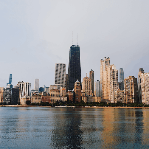 View of Chicago skyline with skyscrapers reflecting on calm lake water under cloudy sky.