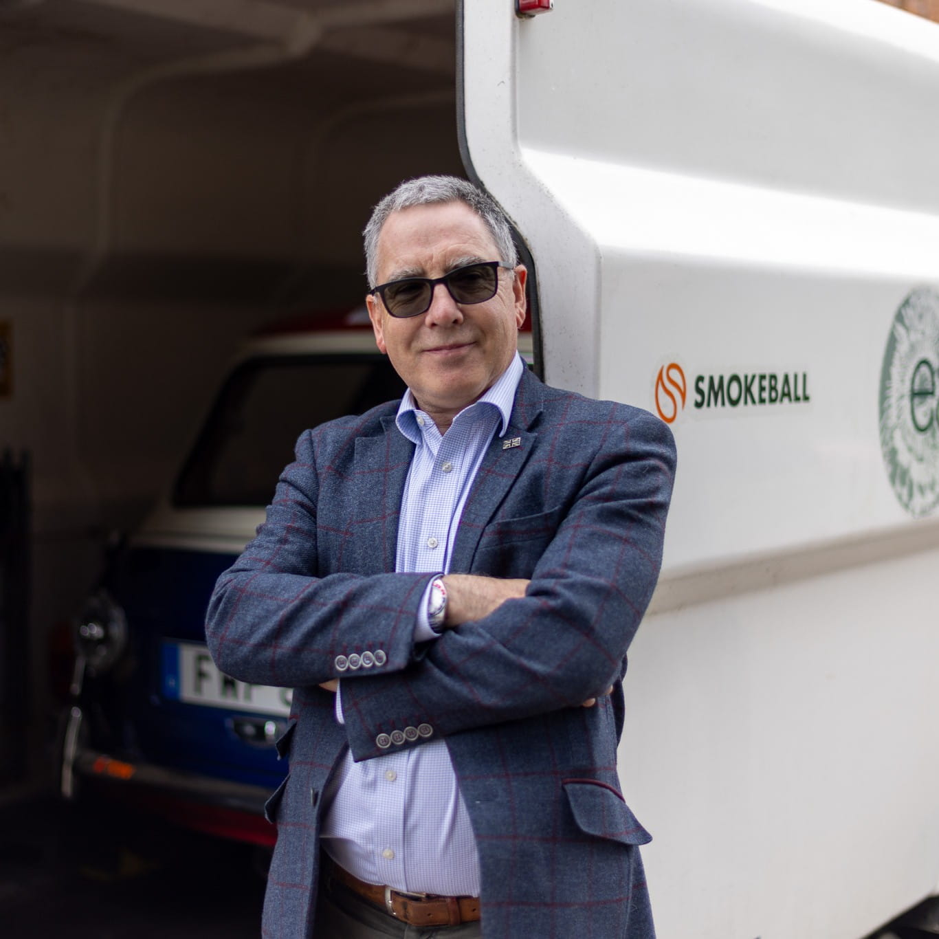 Man wearing sunglasses and blazer stands with arms crossed near a white vehicle with Smokeball logo.