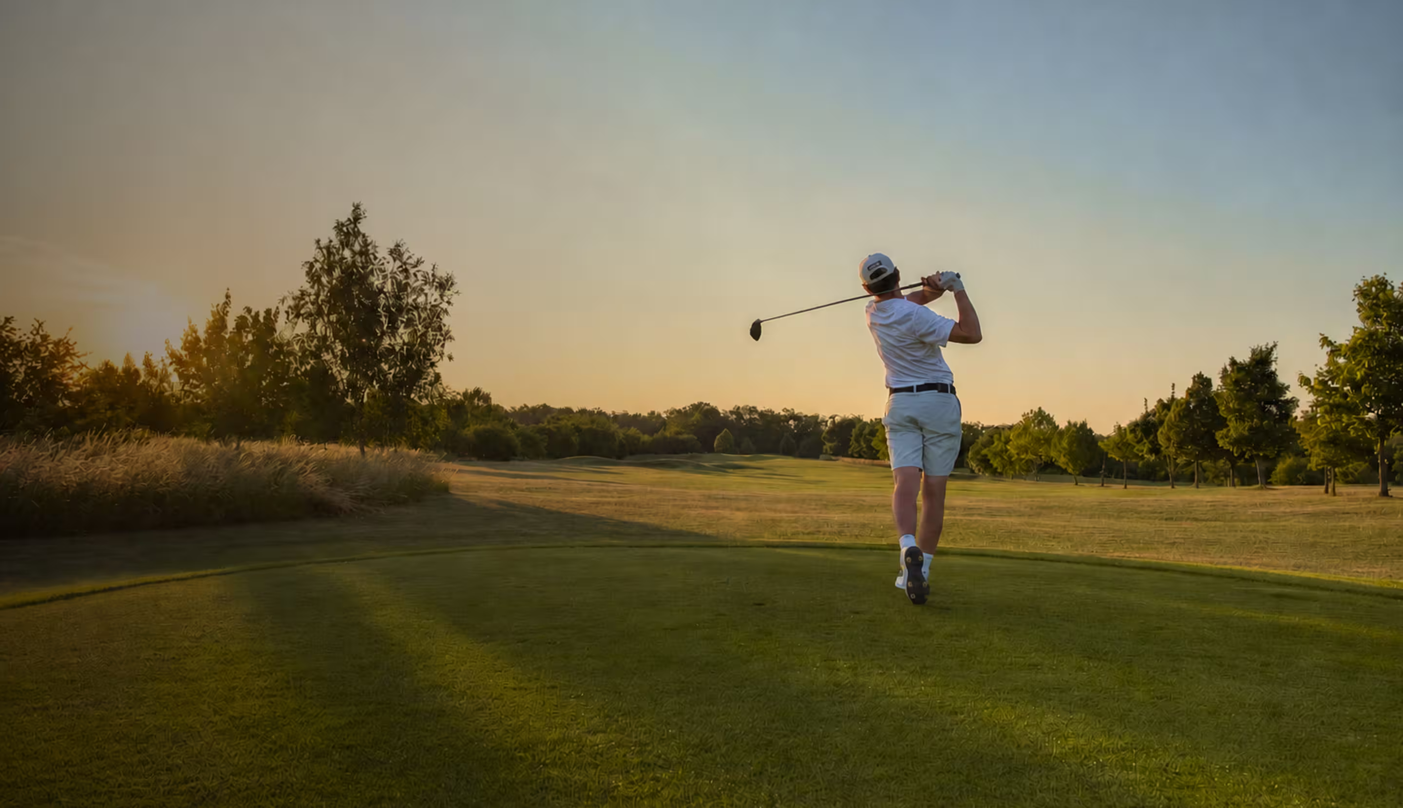 Golfer in white swinging club on a green course at sunset with trees in the background.