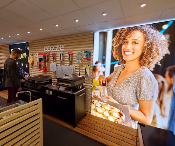 Kvinde med krøllet hår smilende med bageudstyr og pizza baggrund i Cozze-butik.