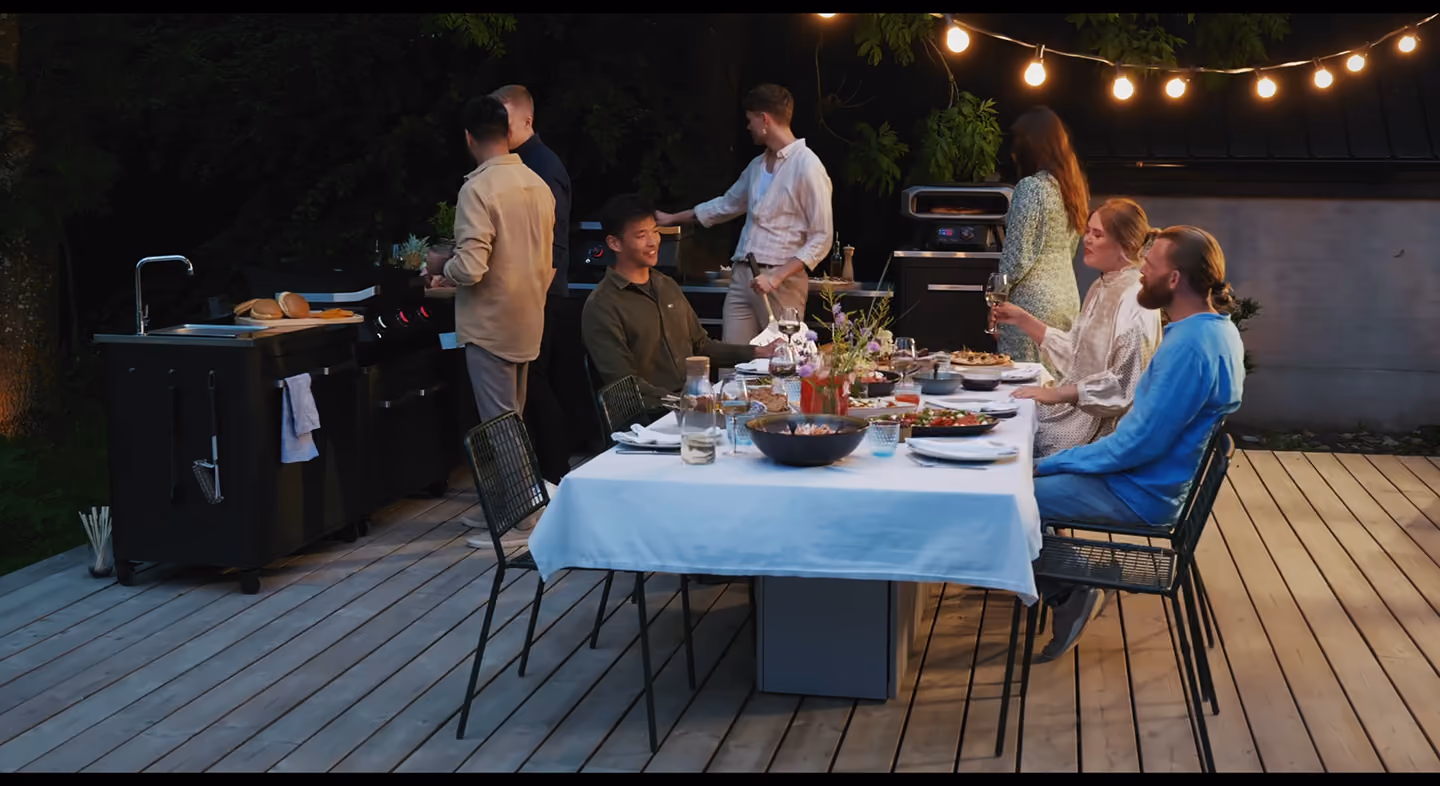 Seks personer nyter en middag utendørs med mat og vin ved et opplyst bord på en terrasse.