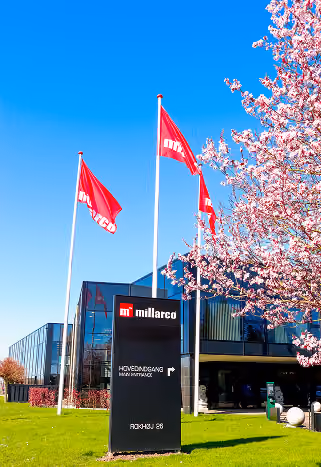 Millarco hovedindgangsskilt med flagsøjler og blomstrende træ foran moderne kontorbygning.