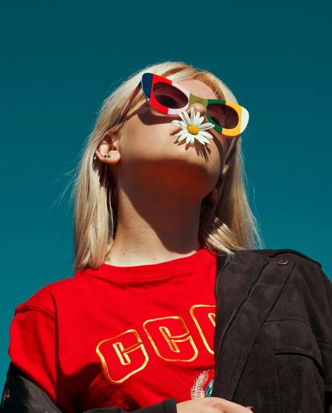 Blonde woman wearing colorful sunglasses and a red CCCP shirt holding a daisy in her mouth against a clear blue sky.