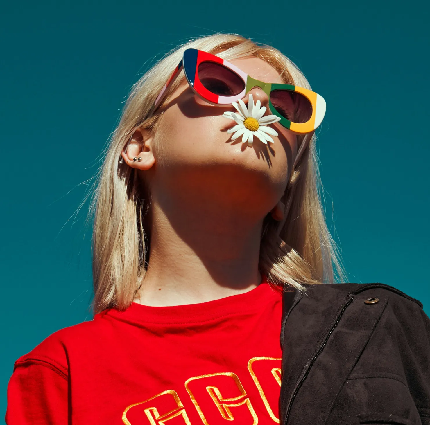Young woman with blonde hair wearing colorful sunglasses and a red shirt with a black jacket over one shoulder holding a daisy flower in her mouth against a clear blue sky.