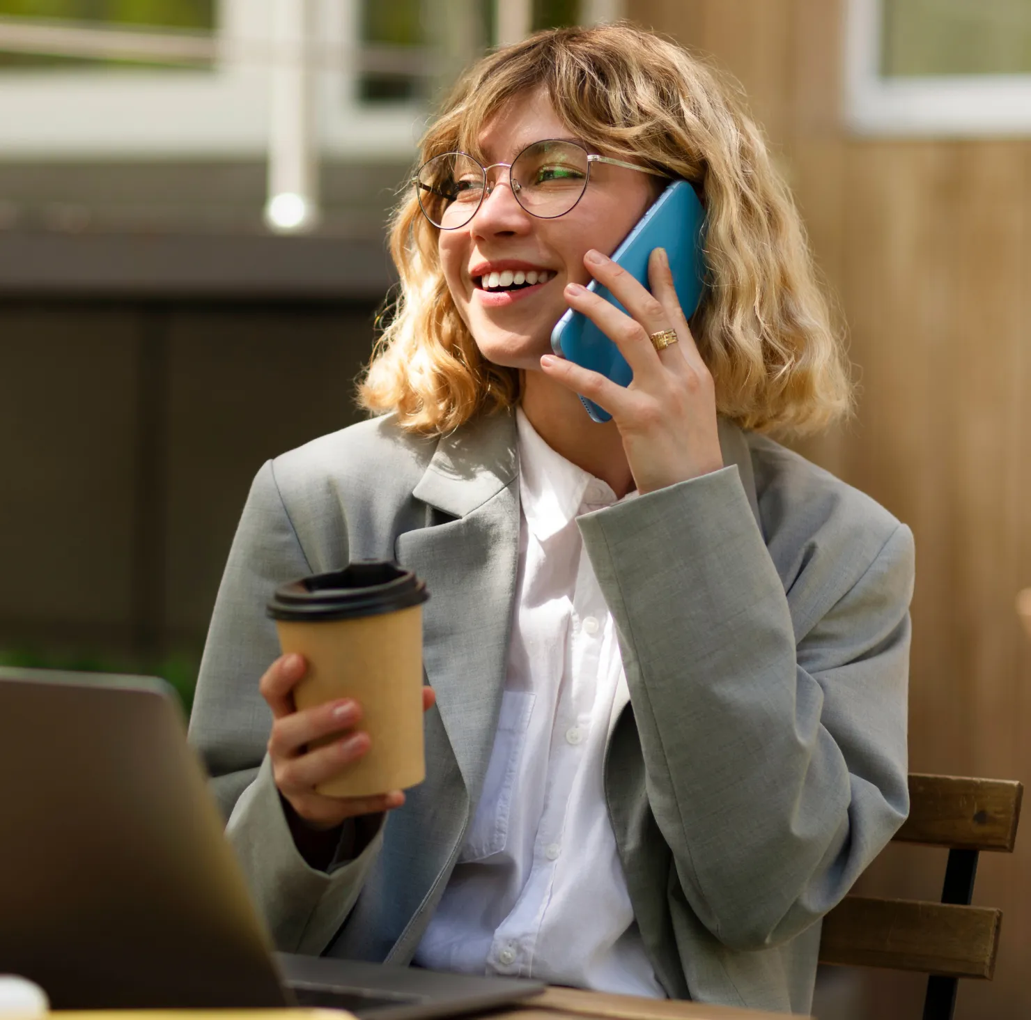 Smiling woman with glasses talking on a blue phone while holding a coffee cup and sitting at an outdoor wooden table with a laptop.