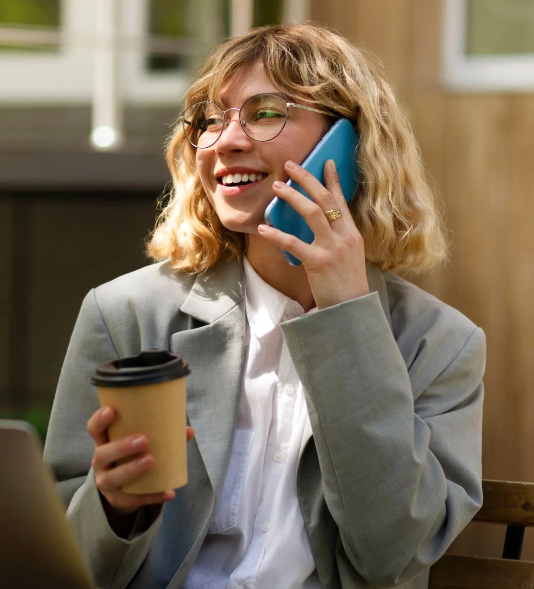 Smiling woman with glasses and blonde curly hair talking on a blue smartphone while holding a coffee cup.