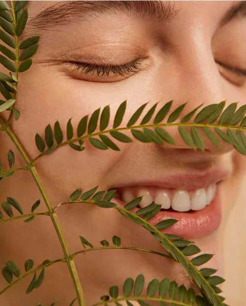 Close-up of a smiling person with closed eyes behind a green leafy branch.