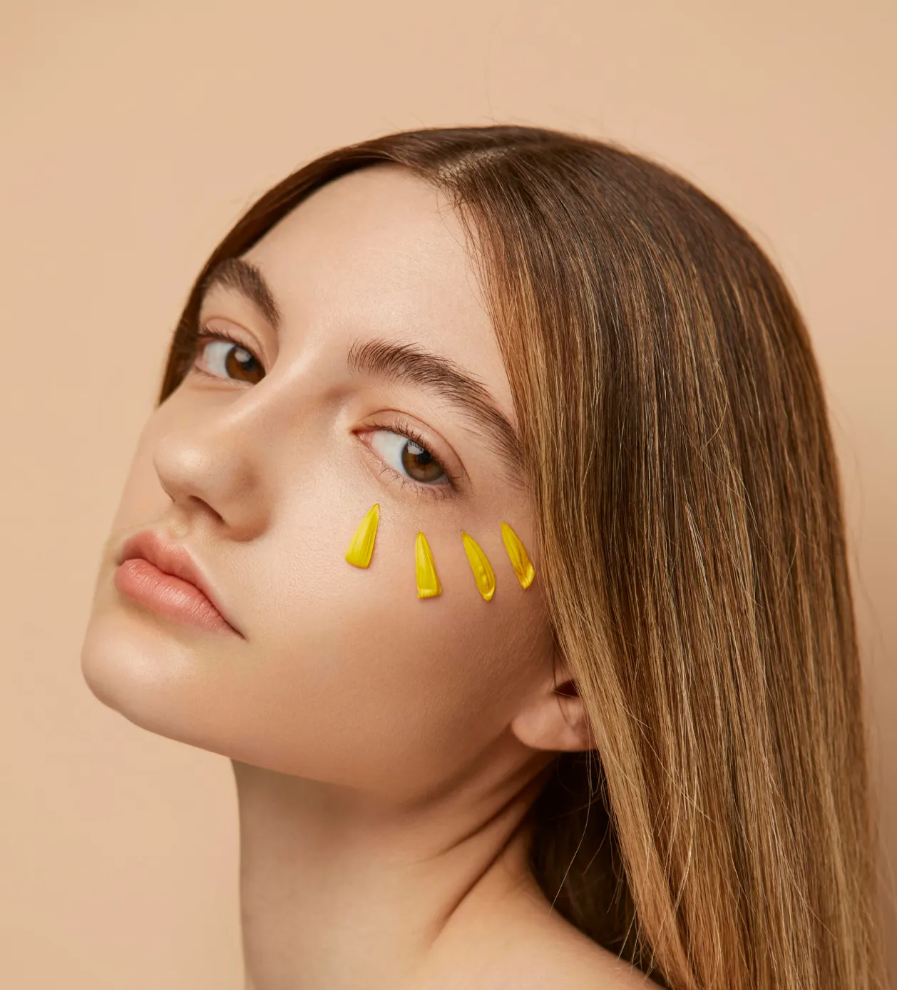 Close-up of a woman with straight light brown hair and yellow flower petals arranged beneath her right eye on beige background.