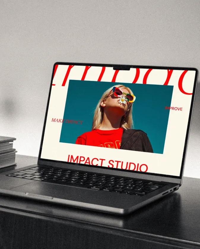 Laptop on a dark desk displaying a website with a photo of a woman wearing colorful sunglasses and holding a daisy flower near her nose, with text 'IMPACT STUDIO' below.