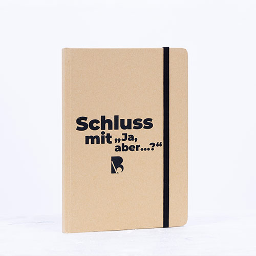 Beige Notizbuch mit schwarzem Gummiband und der Aufschrift „Schluss mit ‚Ja, aber...?‘“ auf der Vorderseite.