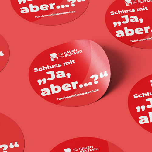 Rote runde Aufkleber mit dem Text „Schluss mit ‚Ja, aber...?‘“ und dem Logo sowie Schriftzug ‚für BAUEN im BESTAND‘ auf rotem Hintergrund verteilt.