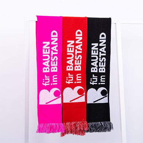 Drei Schals in Pink, Rot und Schwarz hängen nebeneinander an einer weißen Stange, jeder mit weißem Text und Logo, der Text lautet „für BAUEN im BESTAND“.