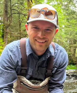 Jeremiah in a cap and sunglasses, crouching by a forest stream