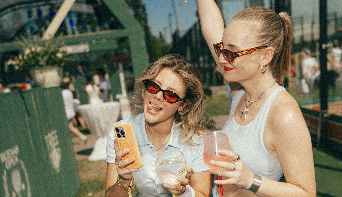 Two guests in sunglasses holding drinks in front of a green padel court