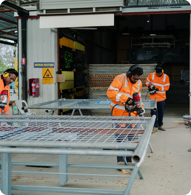 Workers in orange safety gear drilling metal grids inside a warehouse repair section.