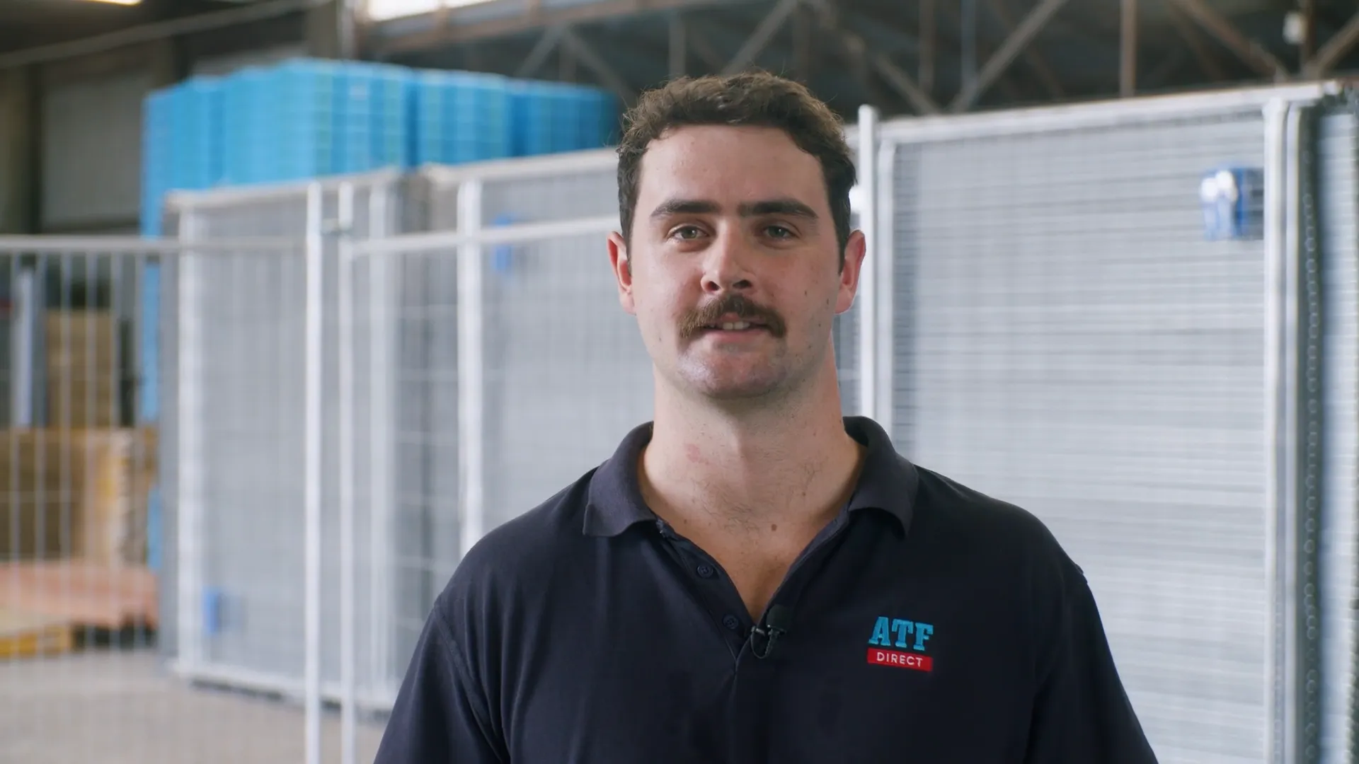 Man with mustache in black ATF Direct polo shirt standing in industrial warehouse setting.