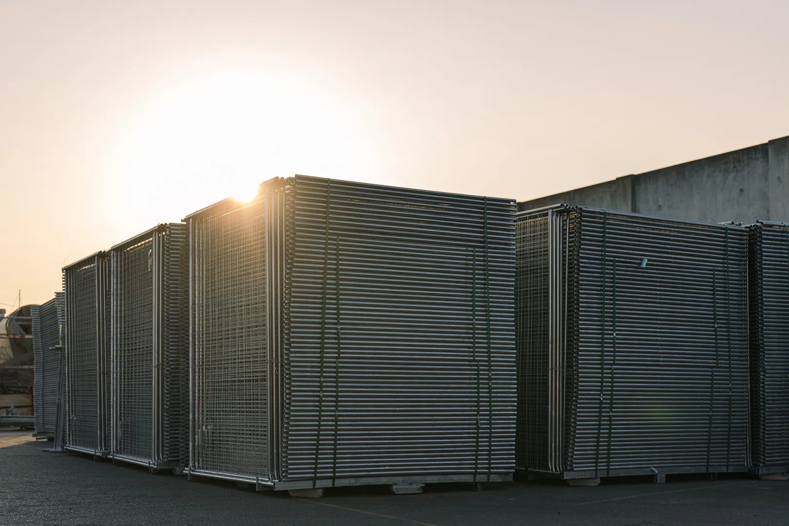 Stacks of metal scaffolding panels tied with green straps, outdoors at sunset near a concrete wall.