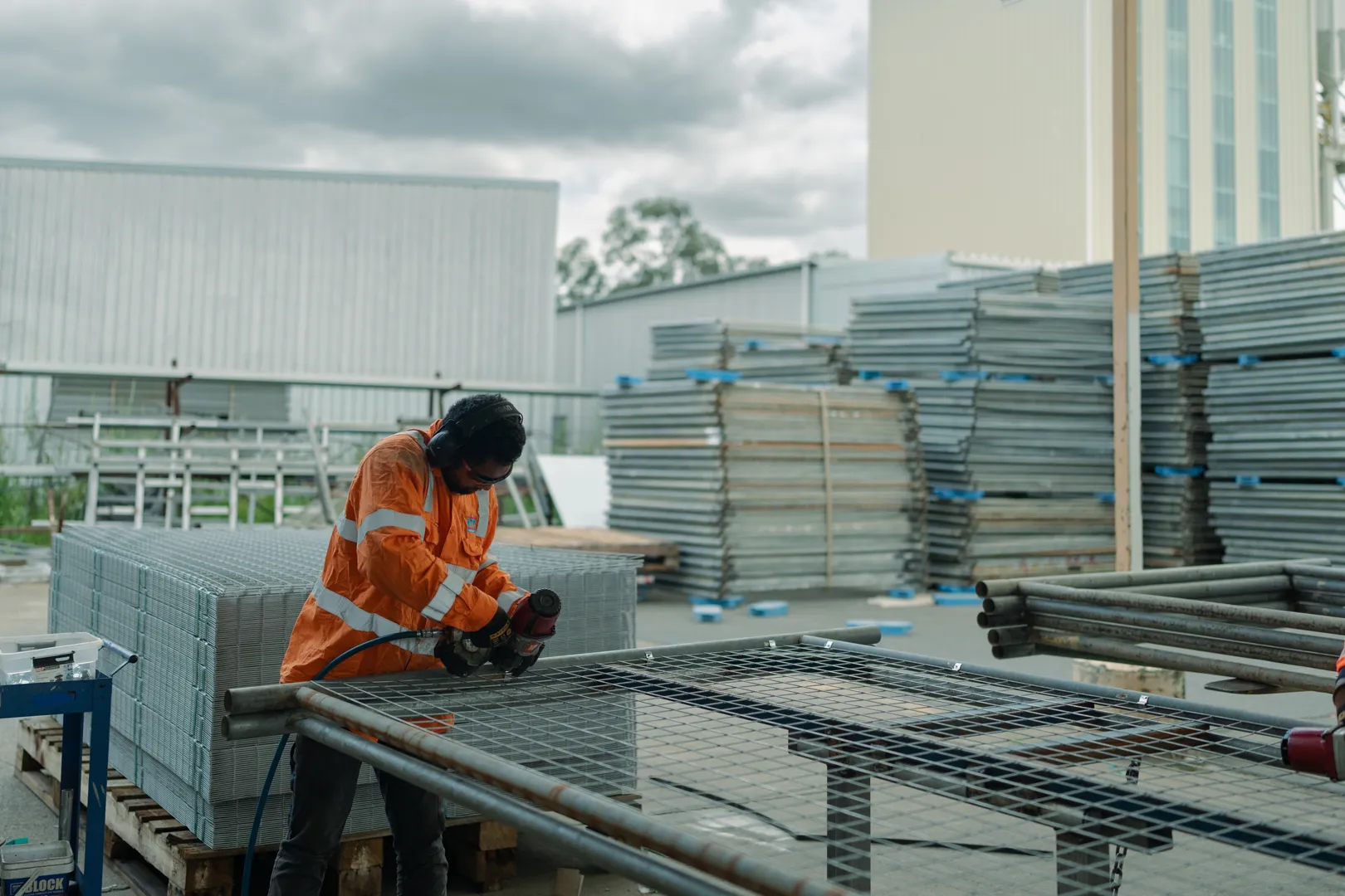 Worker in orange safety gear using a power tool on a metal grid in an industrial yard.