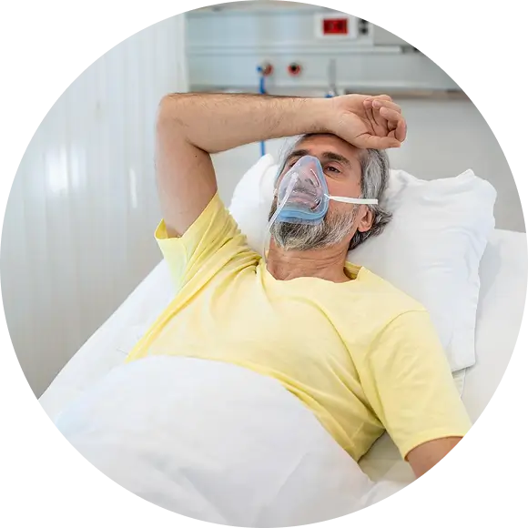Man in yellow shirt lying in hospital bed wearing oxygen mask and resting his arm on his forehead.