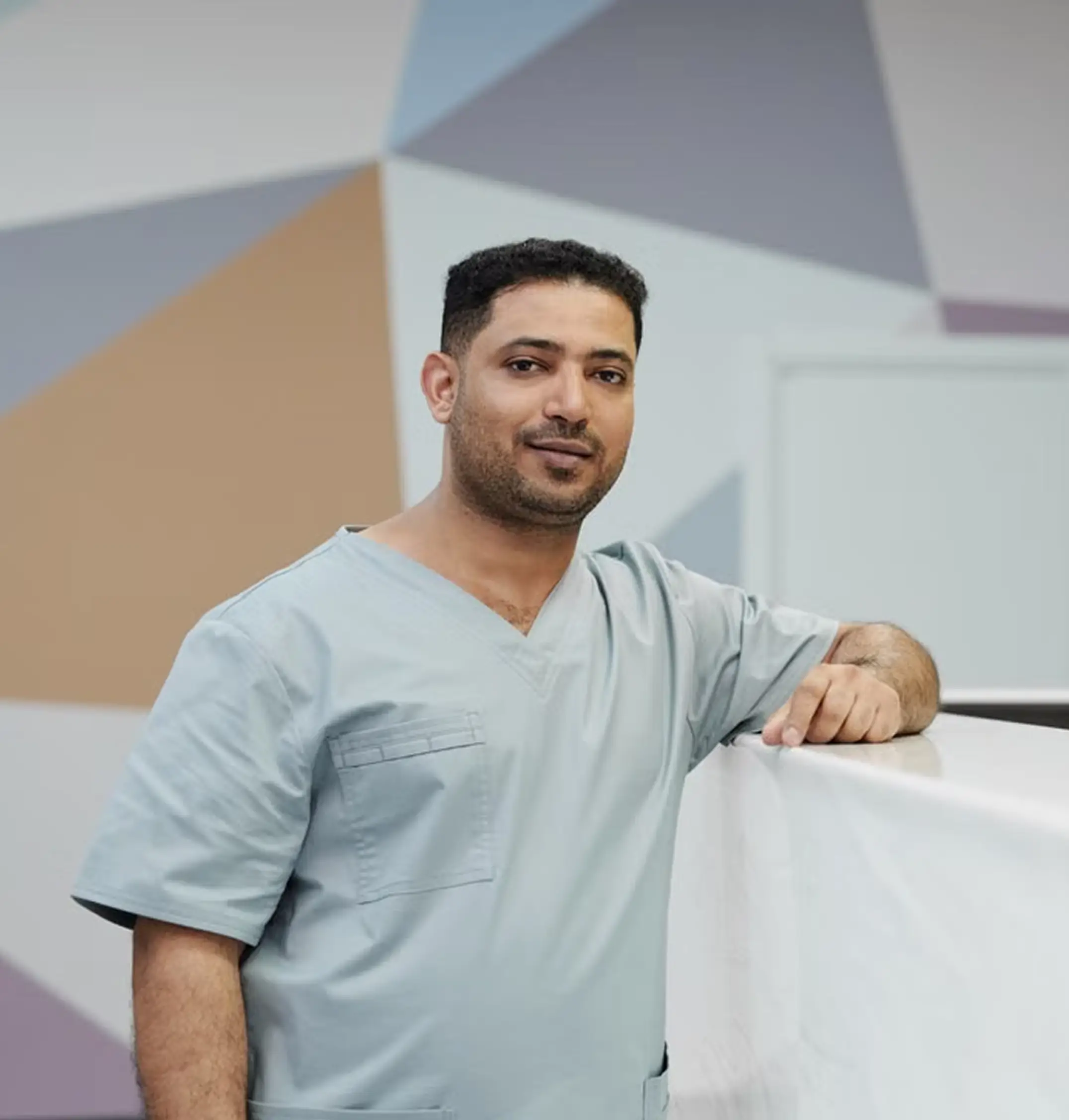 Man in light blue medical scrubs leaning on a white surface with colorful geometric wall behind.