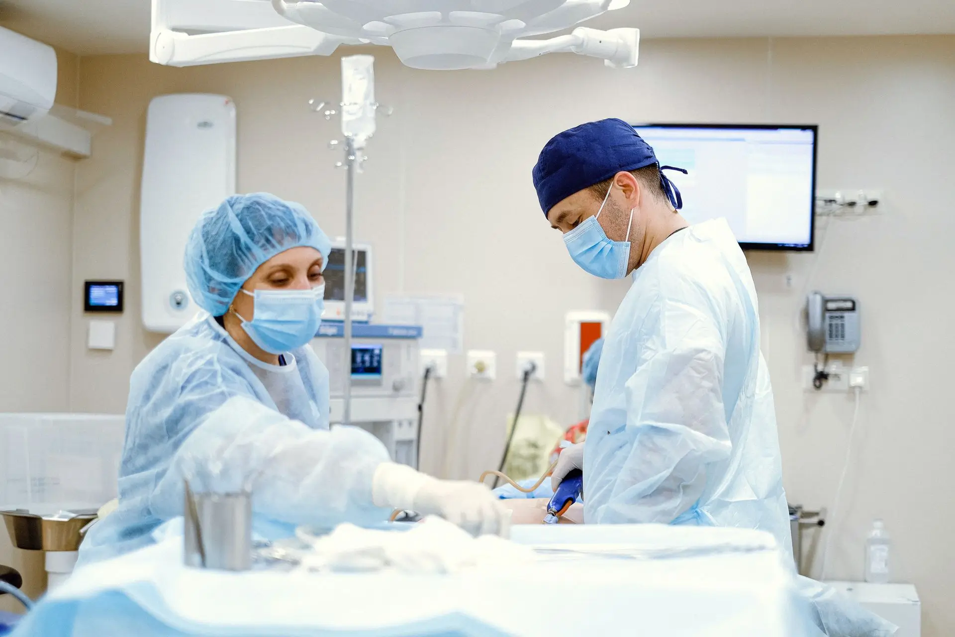Two masked surgeons and a nurse in blue surgical gowns working in a hospital operating room.