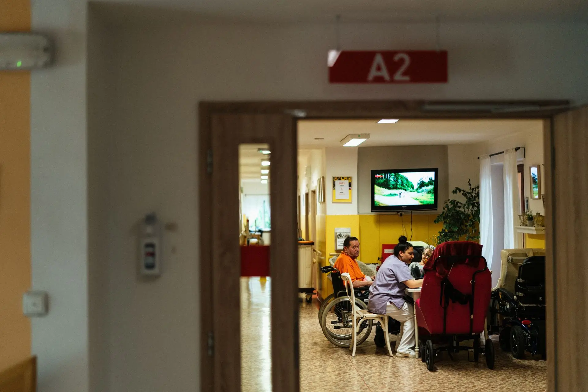 Three people, one in a wheelchair, sitting at a table in a room marked A2 with a TV on the wall.