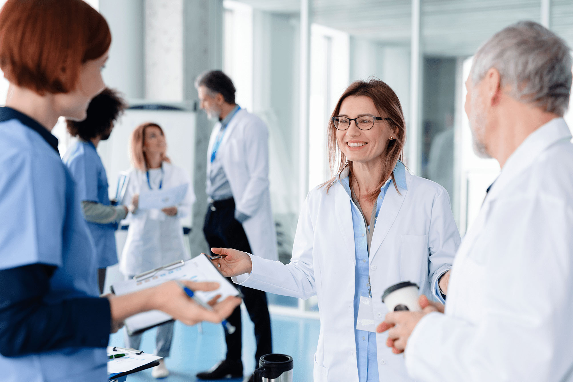 Smiling female doctor with glasses talks to an older male doctor holding a coffee cup.