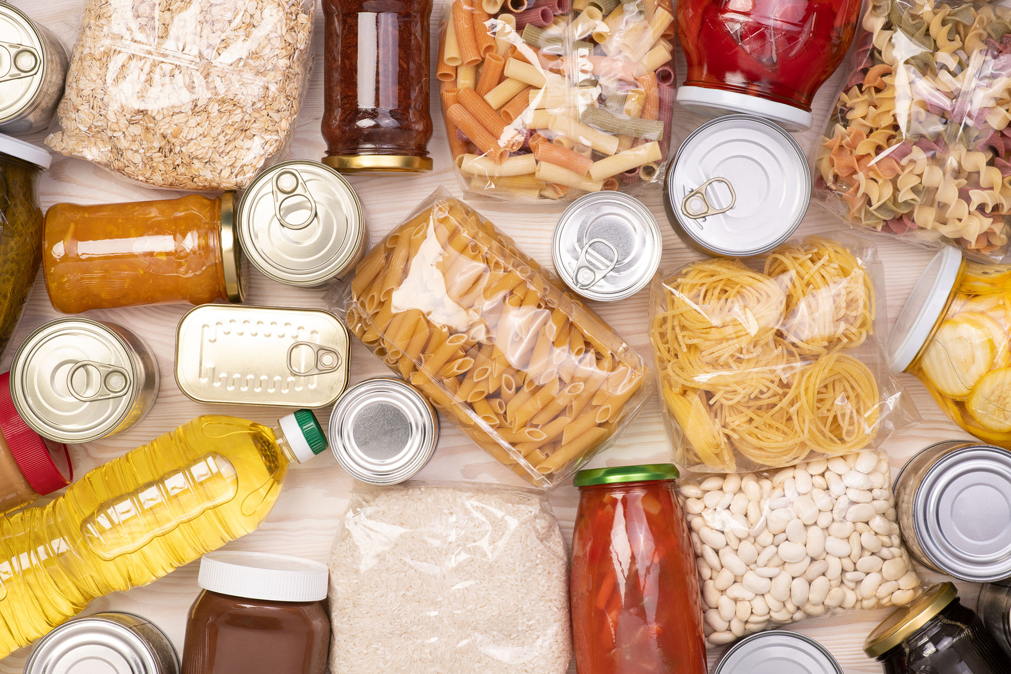 Various packaged dry foods including pasta, rice, canned goods, beans, and jars on a light wooden surface.